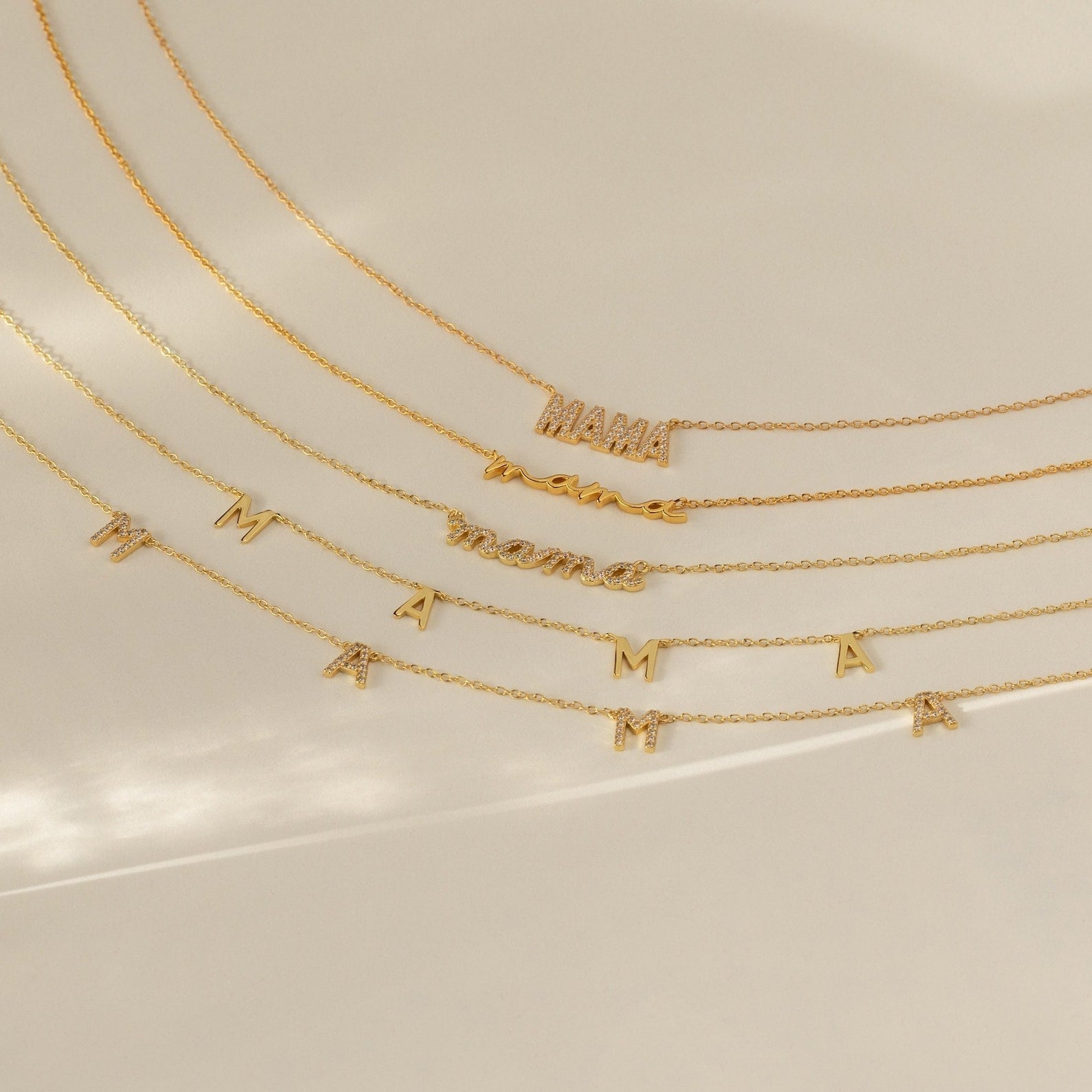 Collection of five gold “mama” necklaces displayed in a cascading layout on a smooth cream background. Designs include pave block letters, cursive script, and individual letter charms with subtle pave accents.