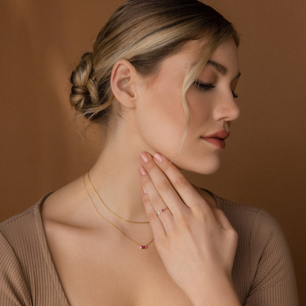 Blonde-haired woman with a low bun, wearing gold jewelry and the Duo Vertical Baguette Ring, gently touches her neck against a brown background.