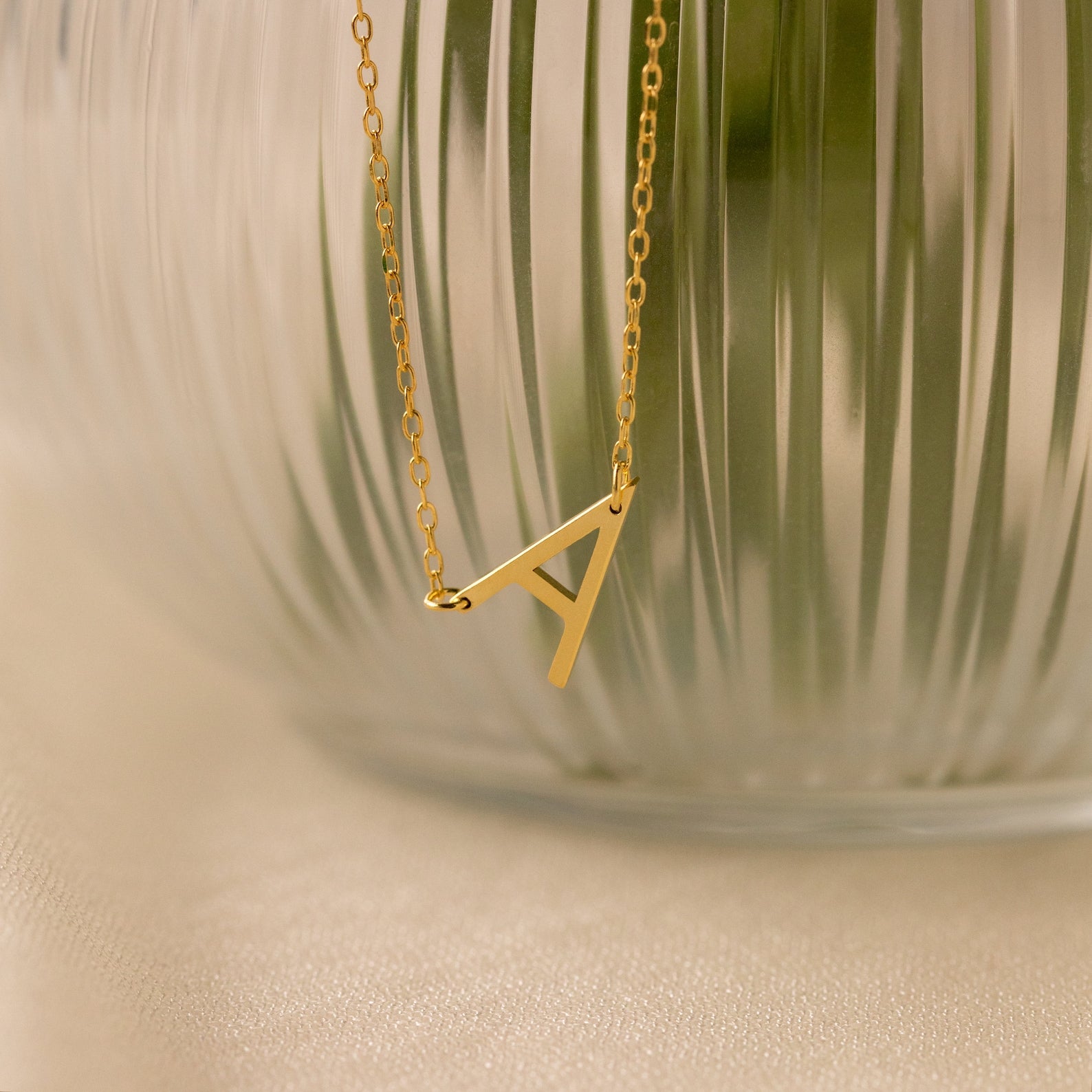 Mini Sideways Initial Necklace featuring an A pendant, displayed in front of a frosted glass vase with vertical lines—an ideal personalized gift.