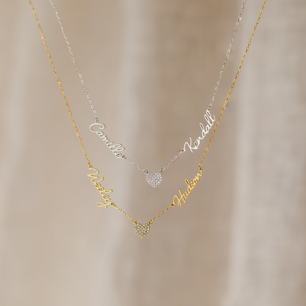 The Grace Name Heart Necklace features two layered name necklaces—one silver, one gold—each personalized in cursive and accented with a sparkling pavé heart charm.