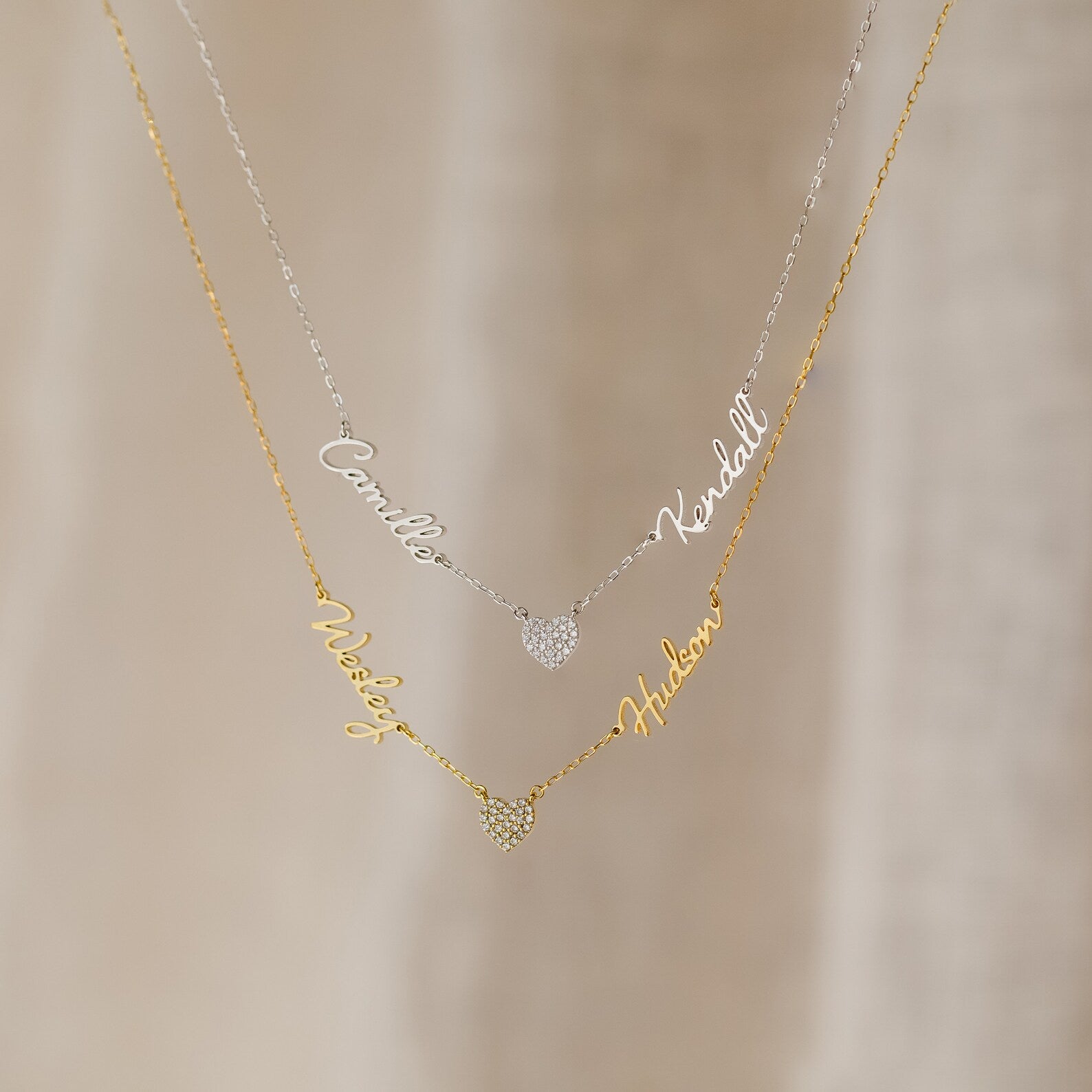 The Grace Name Heart Necklace features two layered name necklaces—one silver, one gold—each personalized in cursive and accented with a sparkling pavé heart charm.