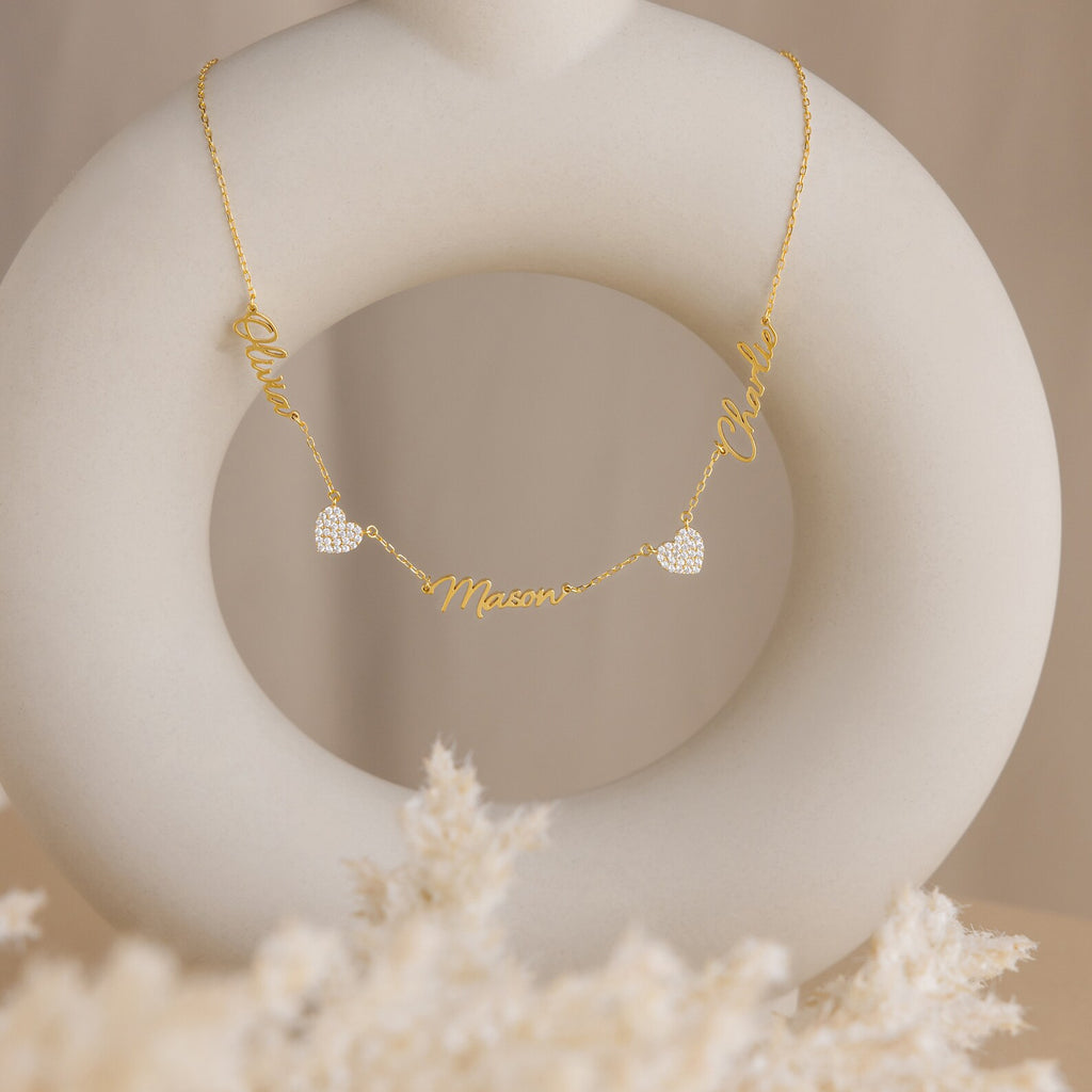 The Grace Name Heart Necklace features three personalized names—Olivia, Mason, and Charlie—with two heart charms, elegantly displayed on a white sculpture.