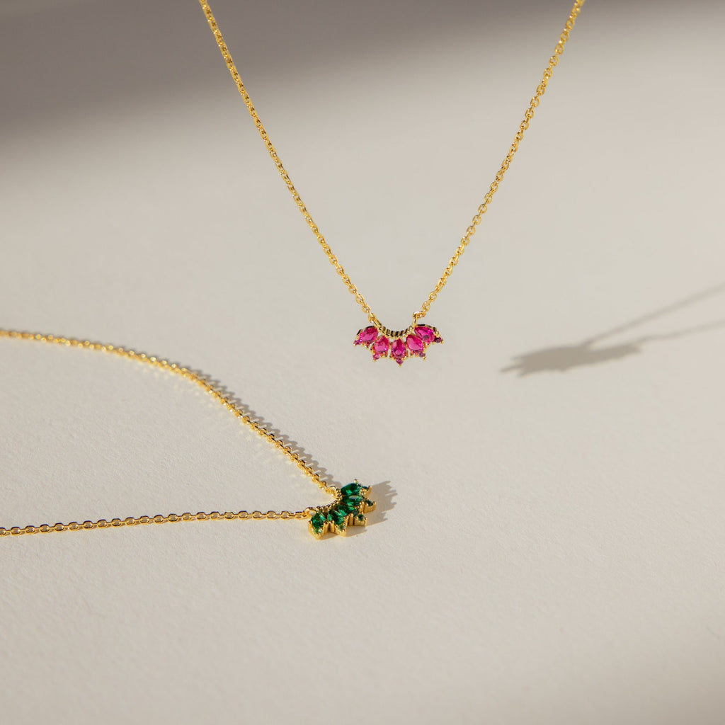 Two delicate gold chain necklaces, one featuring a pink gemstone and the other the Marquise Birthstone Necklace with a green stone, displayed on a light surface.