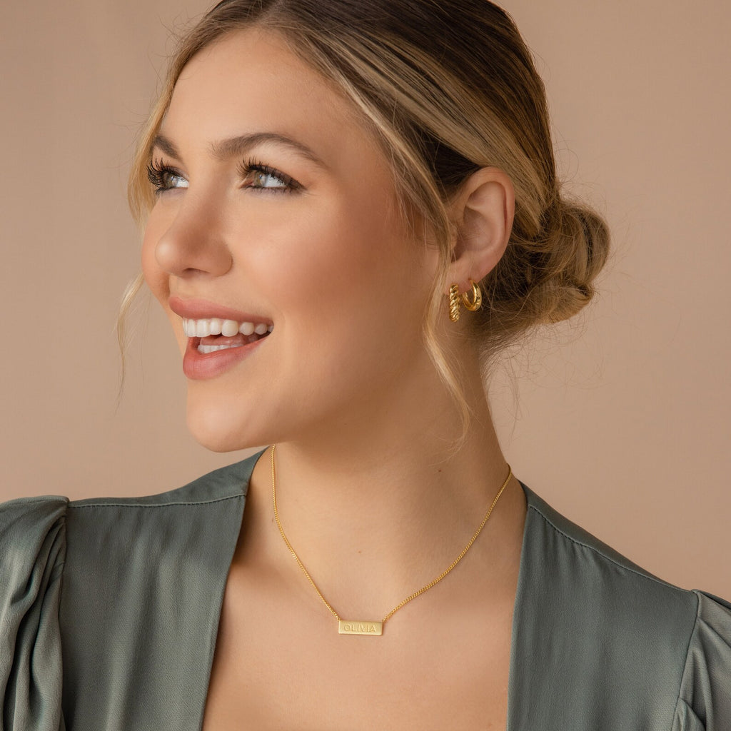 Smiling woman with blonde hair in a bun, wearing gold earrings and a Dainty Bar Name Necklace, looking left.