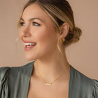 Smiling woman with blonde hair in a bun, wearing gold earrings and a Dainty Bar Name Necklace, looking left.