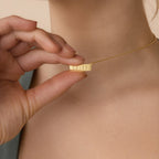 Someone holds the Dainty Bar Name Necklace, a gold personalized bar with engraved vertical lines, close to their neck.