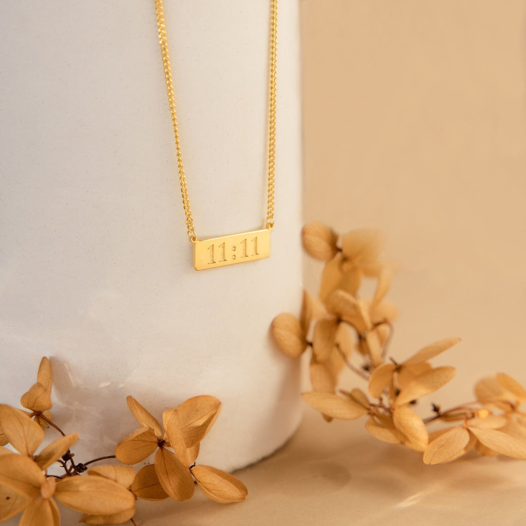 Dainty Bar Name Necklace featuring a rectangular 11:11 pendant in gold, displayed on a white vase with dried beige flowers.