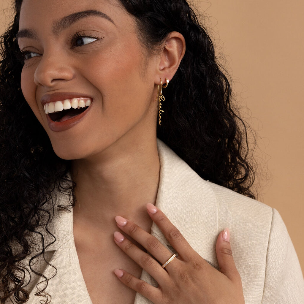 Smiling woman with curly hair wears a cream blazer, a ring, and the Hallie Dangling Name Earrings—showcasing her love for personalized jewelry—as she holds her hand to her chest.