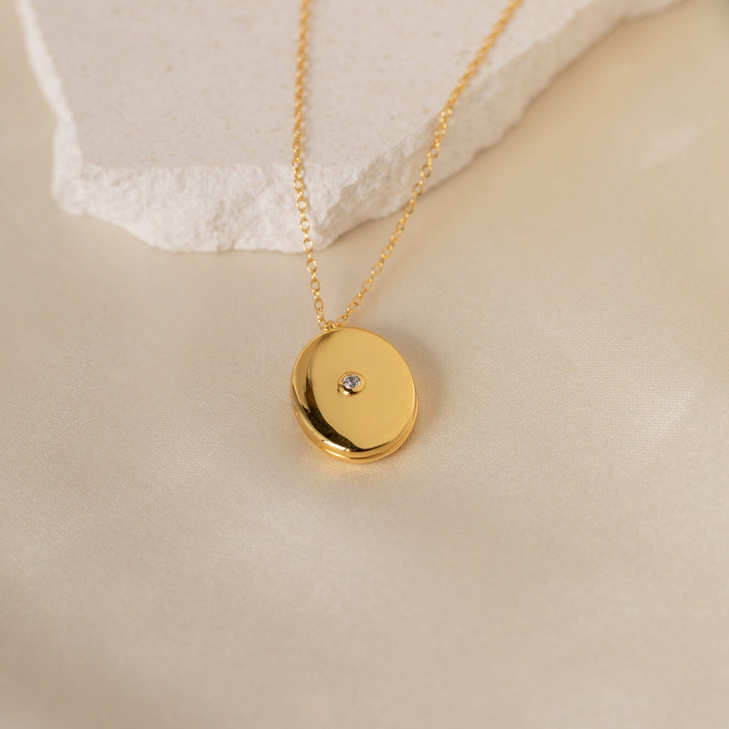 The Eliza Oval Locket Necklace, featuring a small central gemstone, is displayed on a light textured surface—a timeless gift ideal for treasuring precious memories.