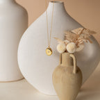 Minimalist scene with white and beige vases, dried flowers, and the Eliza Oval Locket Necklace draped elegantly over a vase—an ideal sentimental gift.