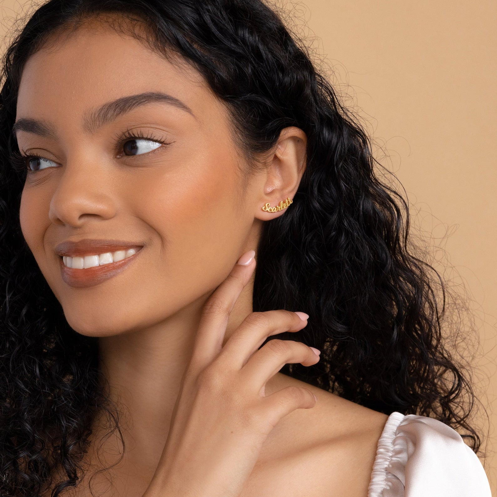 Smiling woman with curly hair wears Mellow Name Earrings and a white top against a beige background.