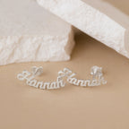 Mellow Name Earrings in silver, spelling "Hannah" in cursive, are displayed on a beige surface with white stones—ideal as personalized earrings or a custom gift.