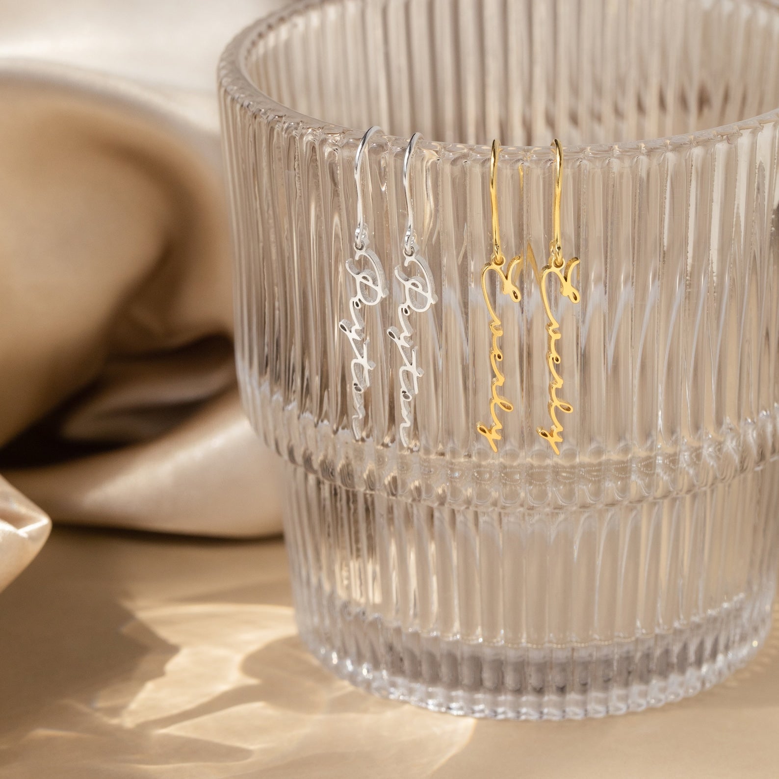 Two pairs of Minimal Dangling Name Earrings—one silver, one gold—are displayed on a ribbed glass cup with a beige satin backdrop, making them the perfect unique bridesmaid gift.
