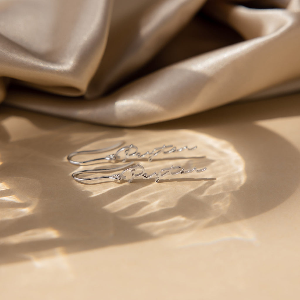 Minimal Dangling Name Earrings are displayed on a beige surface with draped satin fabric in the background—an elegant gift idea for bridesmaids.