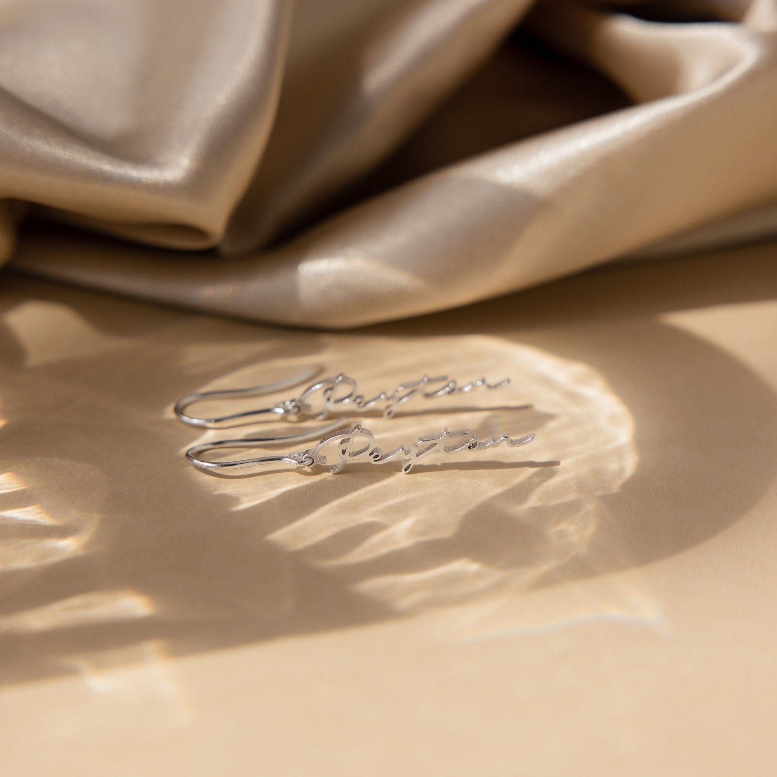 Minimal Dangling Name Earrings are displayed on a beige surface with draped satin fabric in the background—an elegant gift idea for bridesmaids.