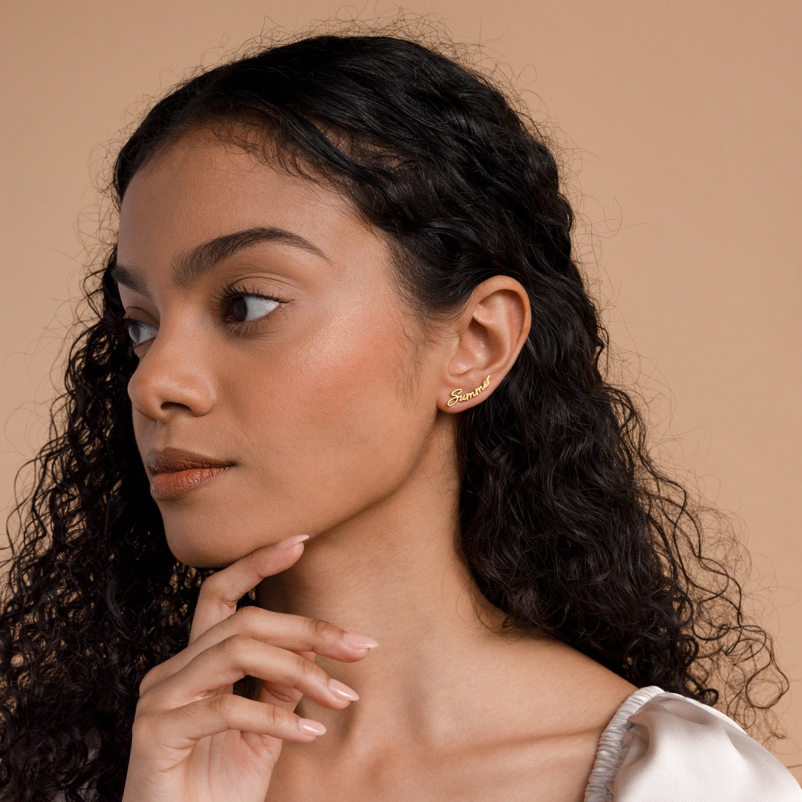 A woman with curly hair wears Grace Name Earrings and a gold ear cuff, gazing to the side with her hand near her chin.