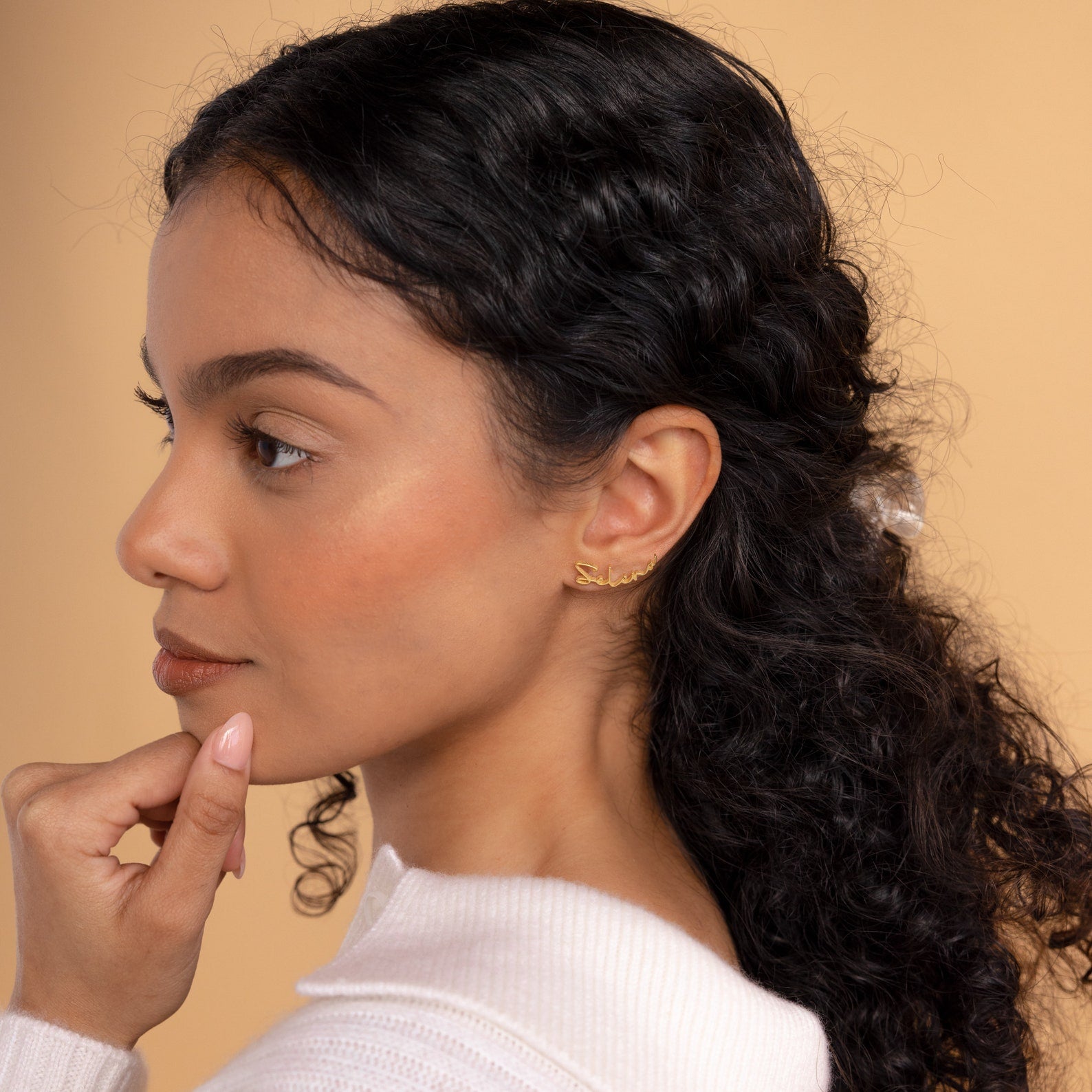 A woman with curly dark hair in a white top wears Summer Name Earrings (925 Sterling Silver), gazing thoughtfully to the side against a beige background, her chin resting on her hand.