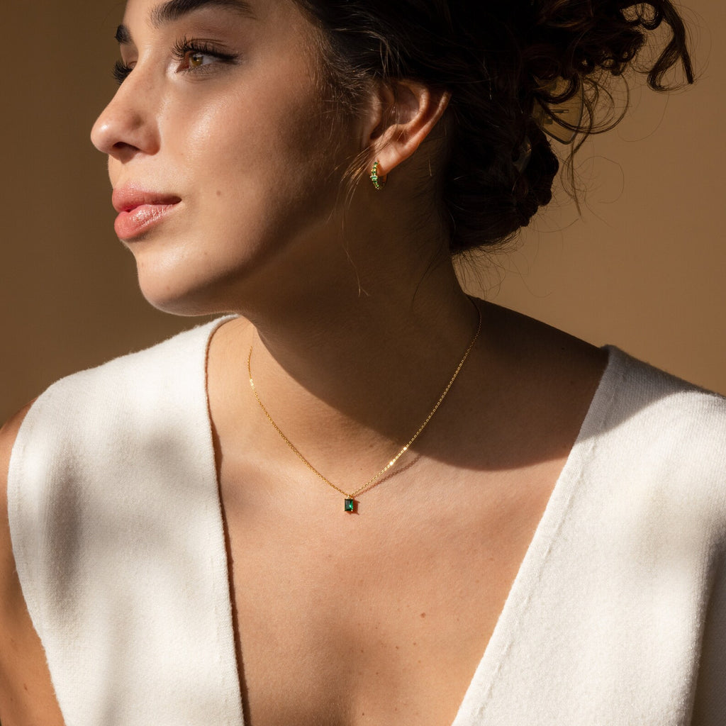 A woman in a white top gazes left, adorned in gold jewelry with green stones—the stunning Maia Emerald Necklace—bathed in soft natural light.