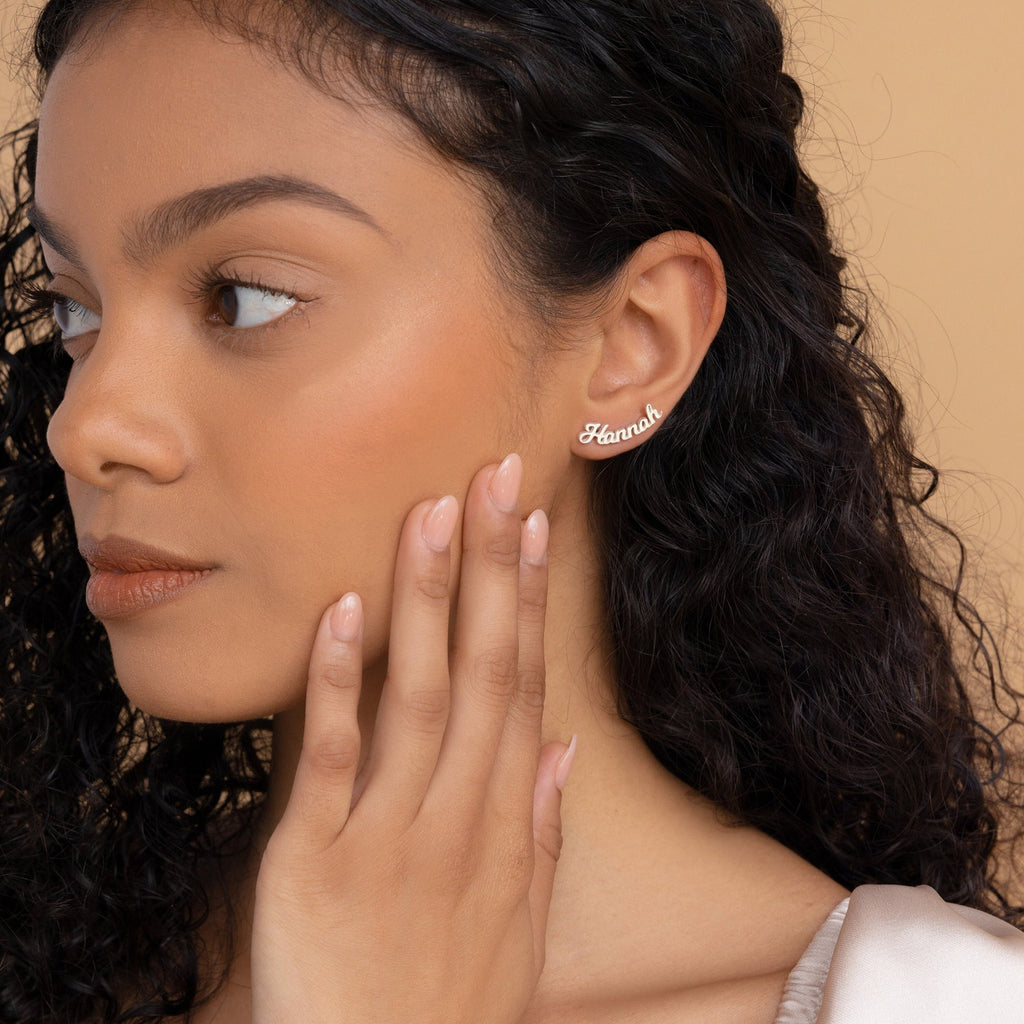 A woman with curly hair touches her face, elegantly showing off Mellow Name Earrings—a personalized custom gift that shines with individuality.