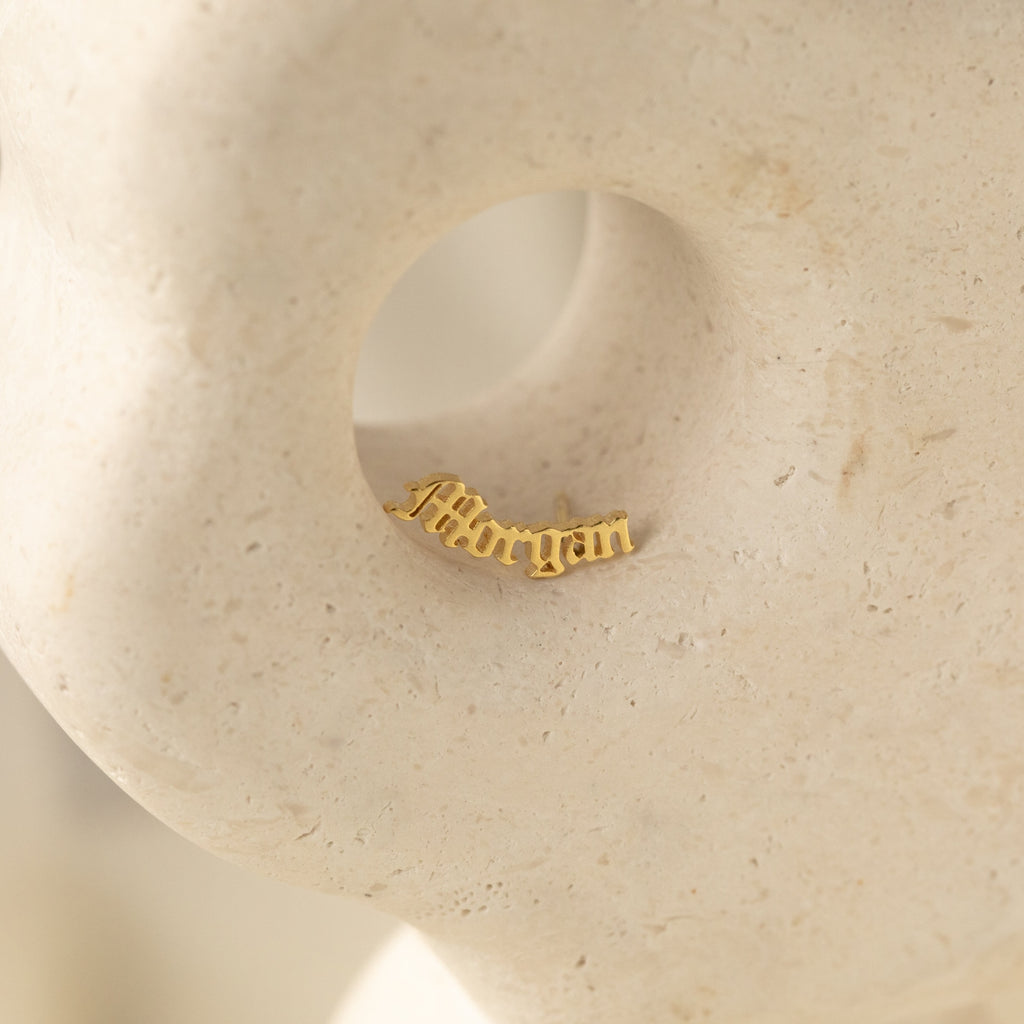Old English Name Earrings displayed in a circular indentation on a textured, light stone surface—a stunning custom gift that pairs beautifully with personalized rings or name stud earrings.