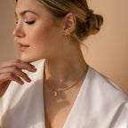 Woman in a white top wearing gold earrings and the Mini Fingerprint Tag Necklace, gazing to the side with her hand near her chin.