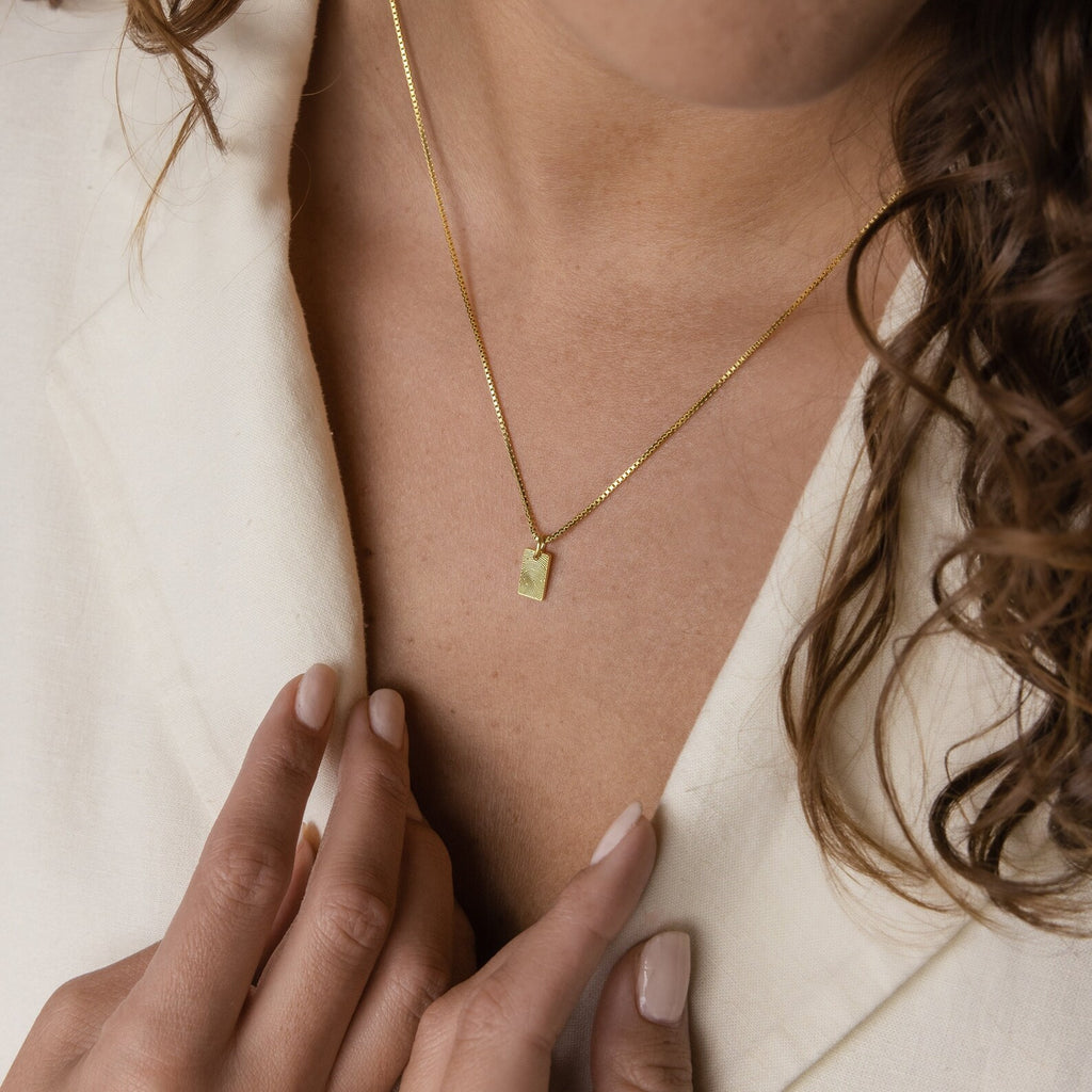 A woman wears the Mini Fingerprint Tag Necklace, a gold rectangular pendant, with a cream top and gently touches her collar—an ideal personalized gift for Mother's Day.
