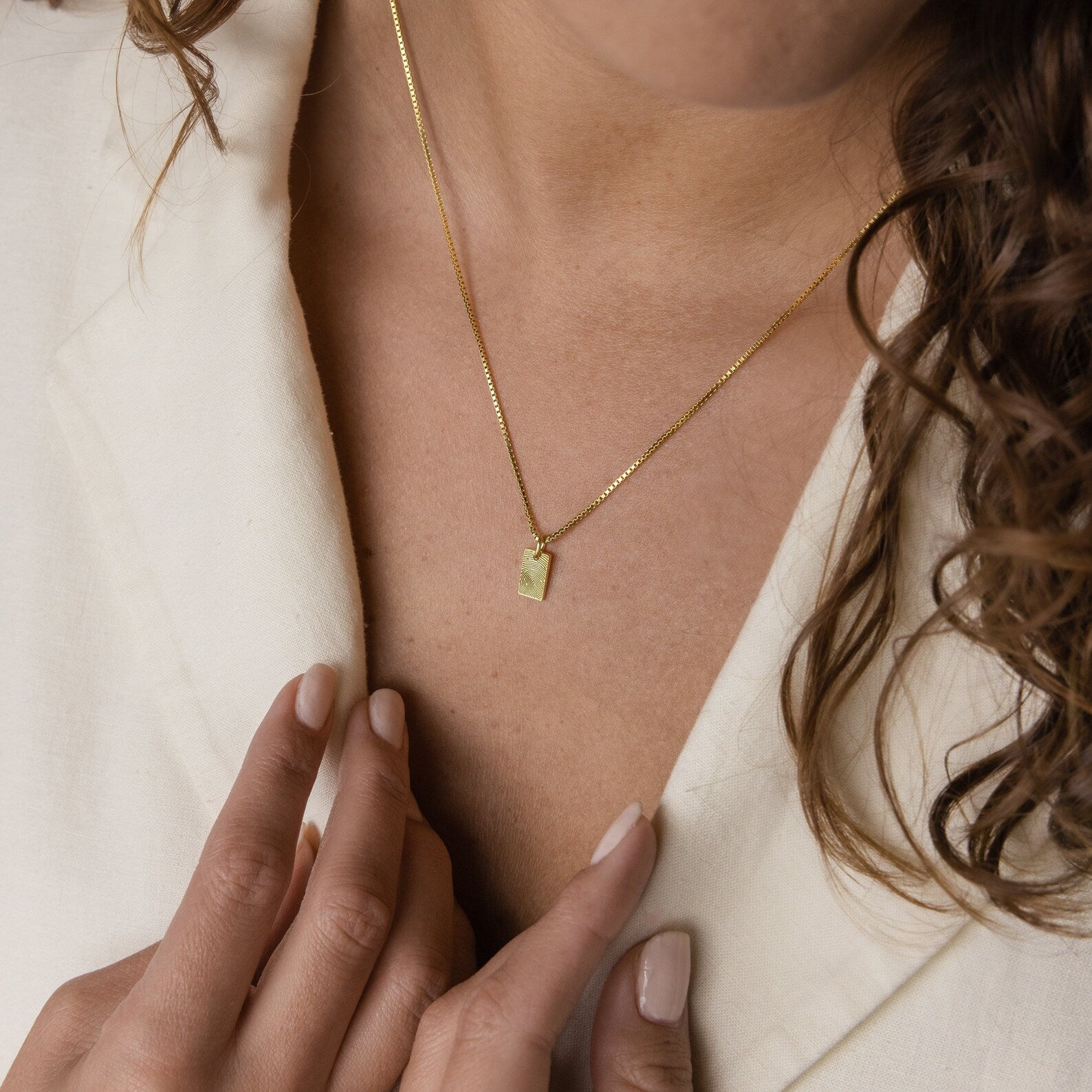 A woman wears the Mini Fingerprint Tag Necklace, a gold rectangular pendant, with a cream top and gently touches her collar—an ideal personalized gift for Mother's Day.