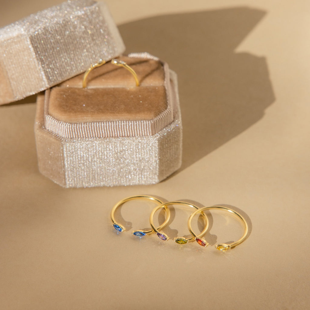 Four gold rings, one being the Duo Marquise Birthstone Ring with colorful gemstones, are arranged next to a beige sparkly ring box on a light tan surface.