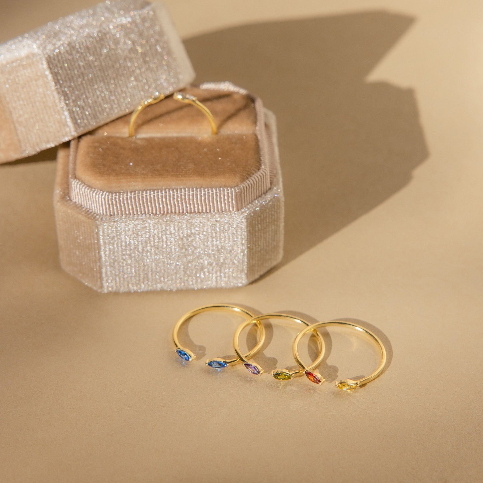 Four gold rings, one being the Duo Marquise Birthstone Ring with colorful gemstones, are arranged next to a beige sparkly ring box on a light tan surface.