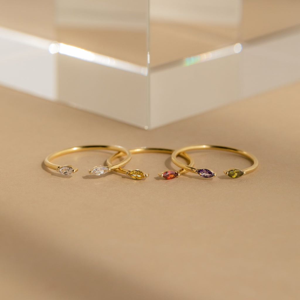 Four delicate gold rings, including the Duo Marquise Birthstone Ring, showcase marquise-cut gemstones in assorted colors and are elegantly displayed on a neutral beige surface.