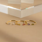Four delicate gold rings, including the Duo Marquise Birthstone Ring, showcase marquise-cut gemstones in assorted colors and are elegantly displayed on a neutral beige surface.