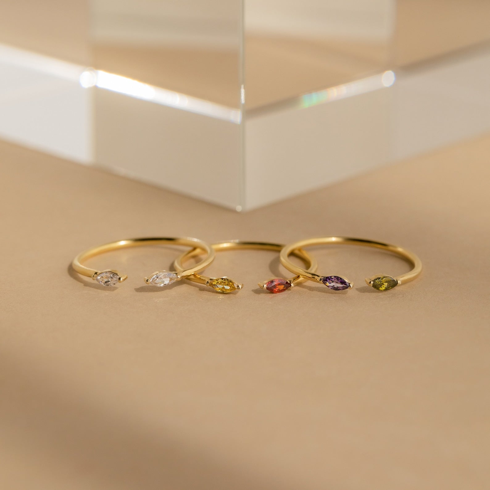Four delicate gold rings, including the Duo Marquise Birthstone Ring, showcase marquise-cut gemstones in assorted colors and are elegantly displayed on a neutral beige surface.