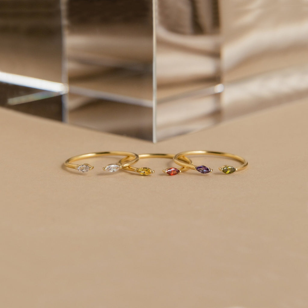Four gold rings, including the Duo Marquise Birthstone Ring, featuring various gemstones are displayed on a beige surface near a clear glass block.