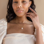 A woman in a cream off-shoulder top gently touches her face, showcasing the Ariel Pearl Necklace—an elegant bridal jewelry choice.