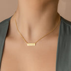 A woman wearing a Dainty Bar Name Necklace with a gold rectangular pendant engraved with the word "helli.
