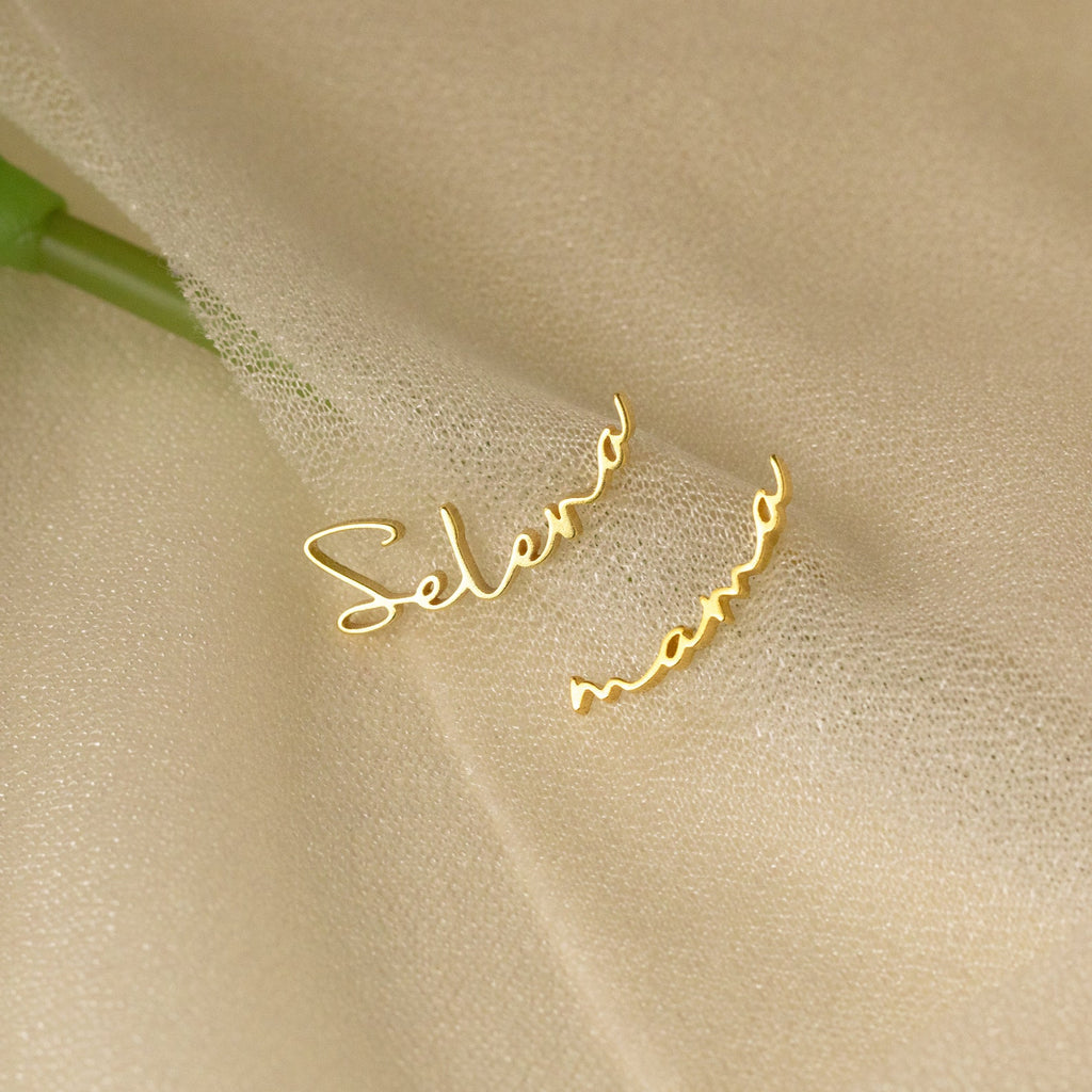 Two Summer Name Earrings with cursive names—one reading Selena and the other mama—rest on a soft beige fabric, showcasing the refined beauty of personalized jewelry.