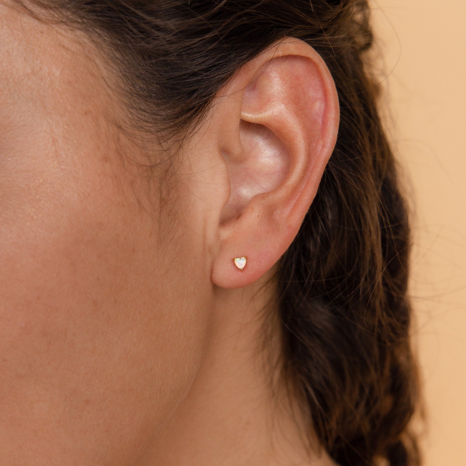 A woman's ear is shown wearing the Opal Heart Studs in gold, highlighting their minimalistic and dainty design against a neutral background.