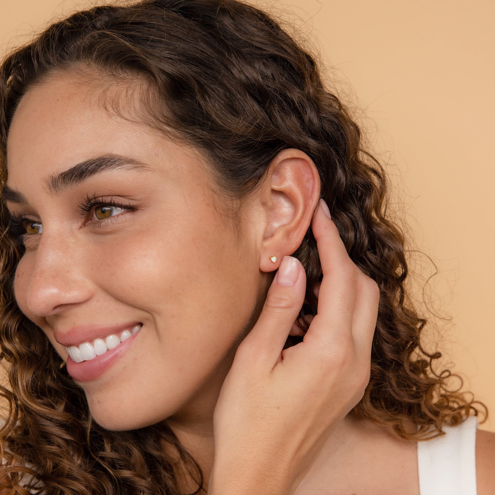 A woman with curly hair smiles and touches her Opal Heart Studs earring, radiating minimalistic glam against a beige background.