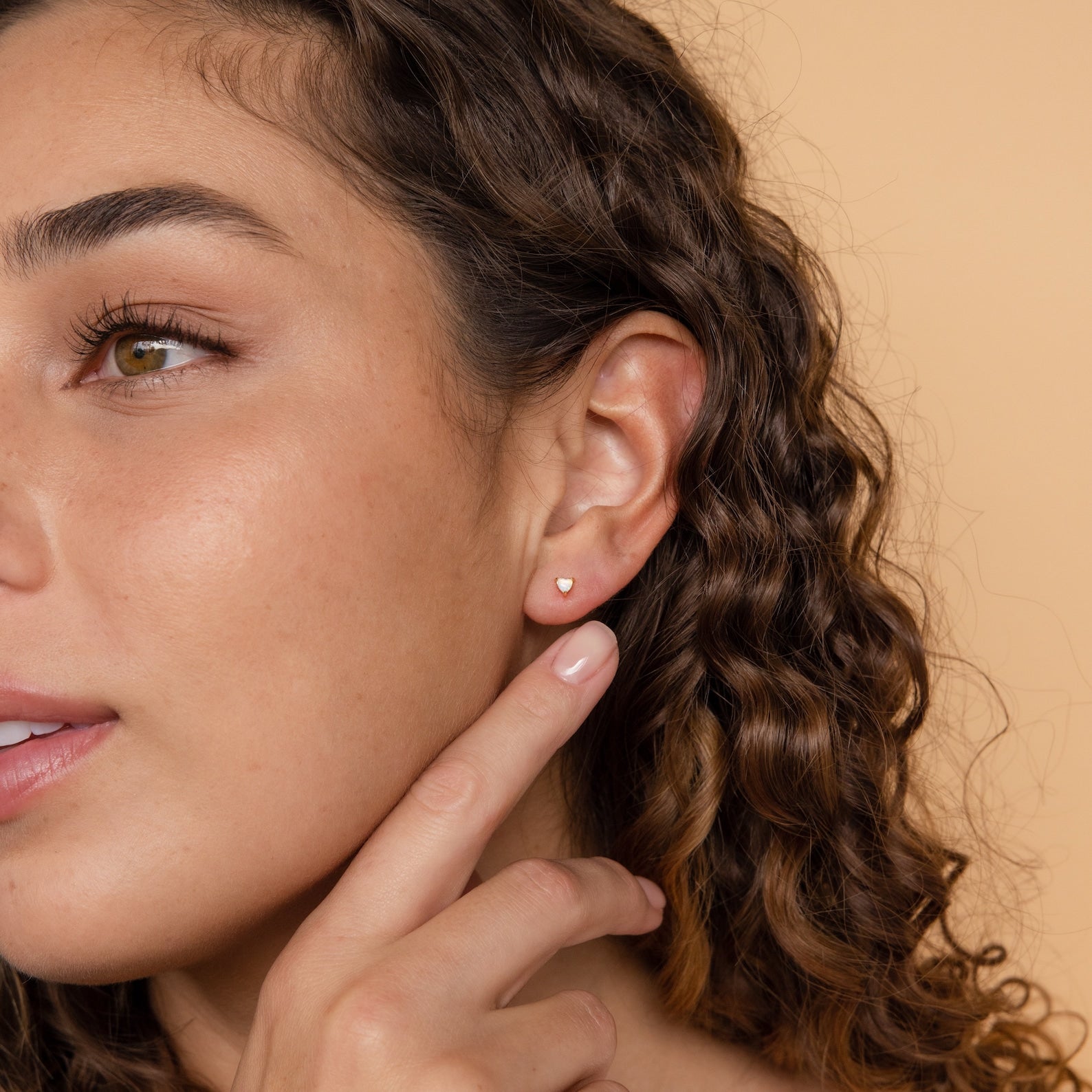 A woman with curly hair points to an Opal Heart Stud in her ear, highlighting the minimalistic glam of these dainty Opal Heart Studs against a beige background.