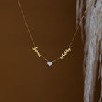 The Mellow Name Heart Necklace features "James" and "Ashley" connected by a small heart charm on a delicate gold chain, displayed against a brown background.