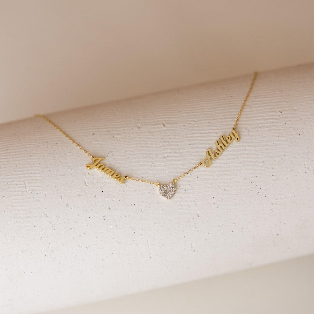 The Mellow Name Heart Necklace features gold personalized names, James and Ashley, connected by a small heart pendant on a dainty chain, displayed on a white textured surface. Perfect for adding a sweet touch to any outfit.