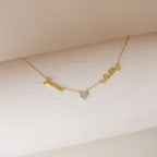 The Mellow Name Heart Necklace features gold personalized names, James and Ashley, connected by a small heart pendant on a dainty chain, displayed on a white textured surface. Perfect for adding a sweet touch to any outfit.