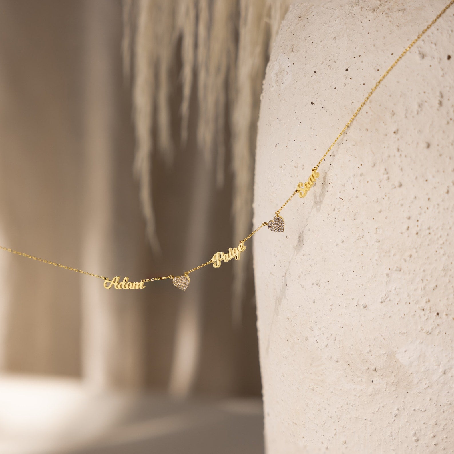 The Mellow Name Heart Necklace features "Adam" and "Paige" in gold, separated by heart charms and displayed on a textured white surface. This dainty chain adds a delicate, personalized touch to any outfit.