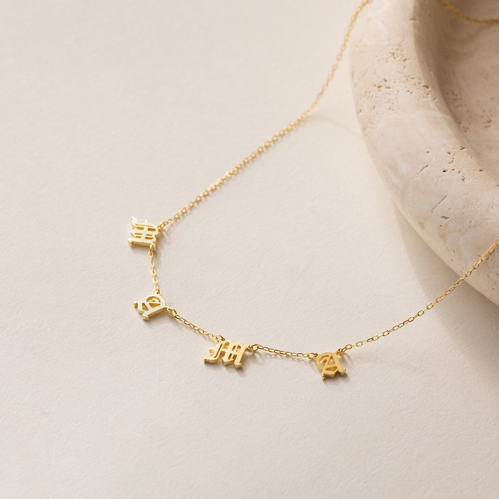Old English Letter Name Necklace with small dangling charms, shown on a light stone surface—ideal for fans of minimalist jewelry with an edgy vibe.