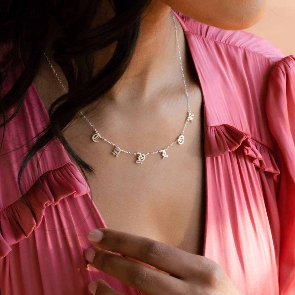 A woman in a pink dress wears the Old English Letter Name Necklace, silver with charms spelling T E S T, adding minimalist jewelry style.
