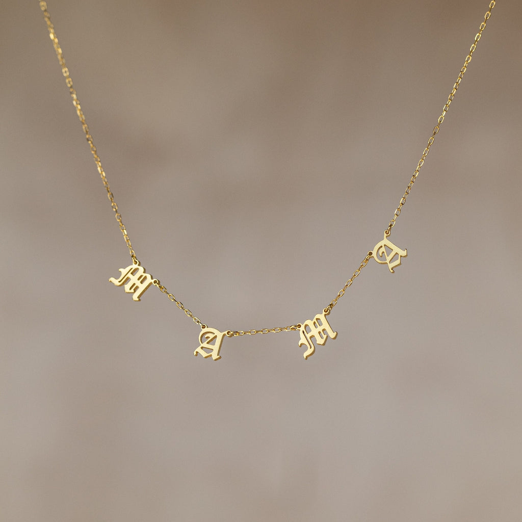 The Old English Letter Name Necklace features dangling letter charms spelling "mama" in a decorative font on a thin gold chain—an ideal minimalist jewelry piece.