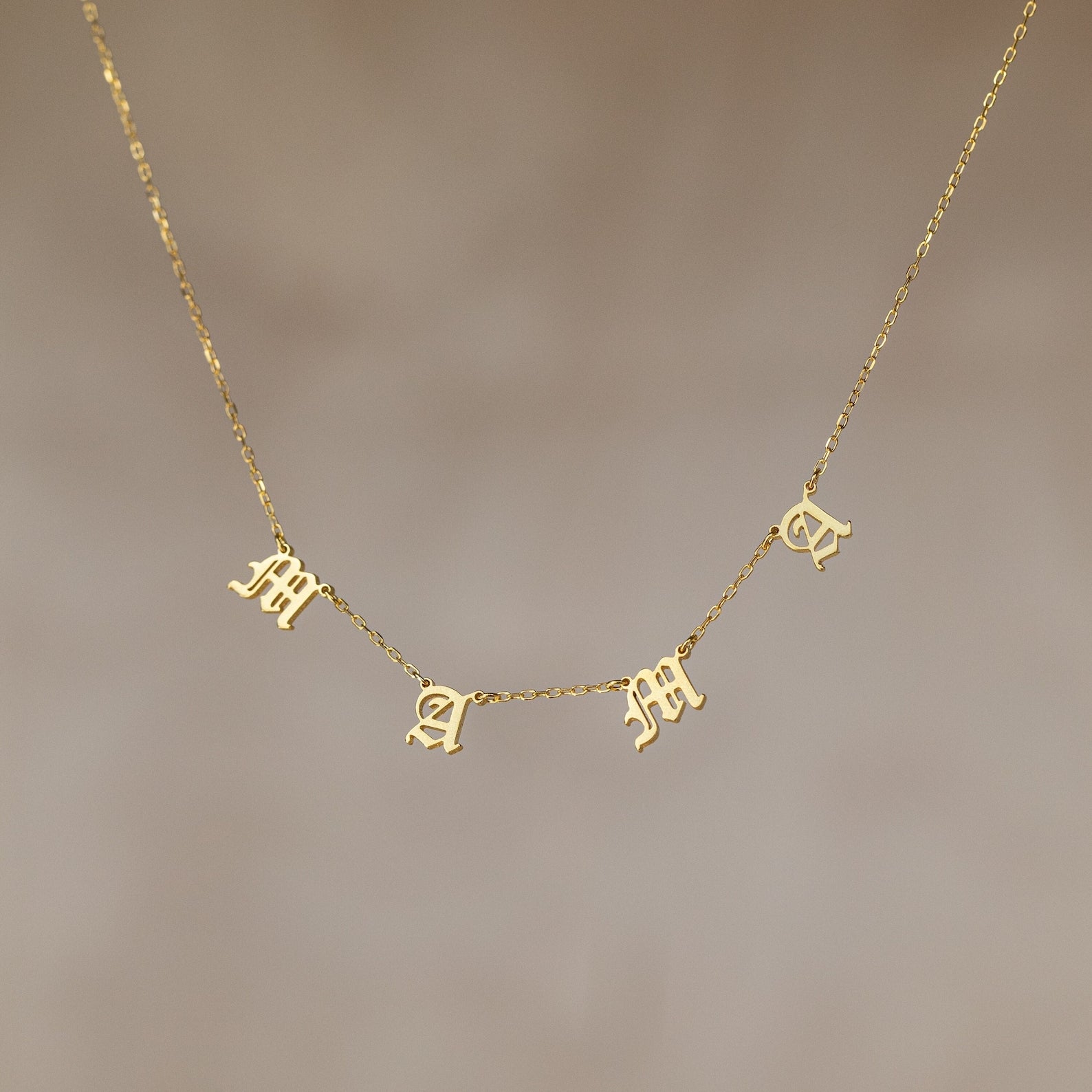 The Old English Letter Name Necklace features dangling letter charms spelling "mama" in a decorative font on a thin gold chain—an ideal minimalist jewelry piece.
