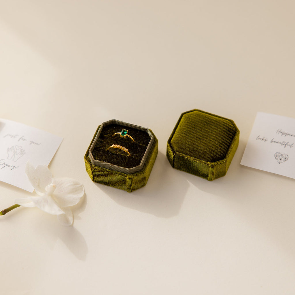 The 2 Slot Velvet Ring Box in elegant green displays a gold ring with emerald inlay, set beside a white flower and cards on a cream surface.