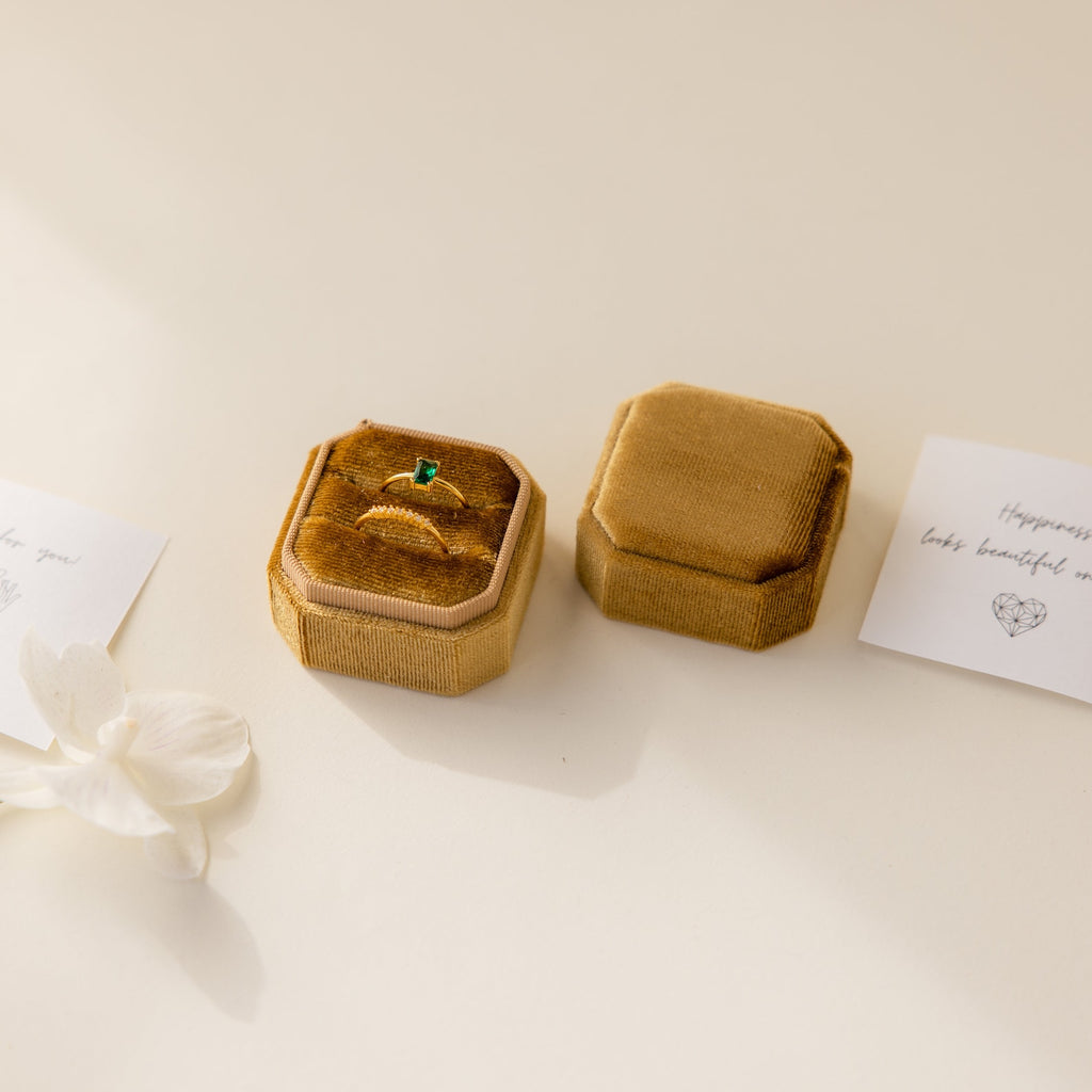 An elegant gold ring with a green stone rests in a 2 Slot Velvet Ring Box on a cream surface, accompanied by handwritten notes and a ring holder.
