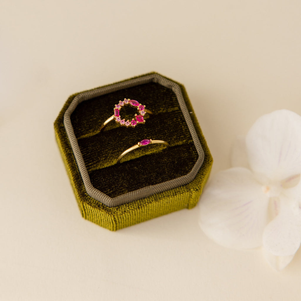 Two gold rings with pink stones sit in a 2 Slot Velvet Ring Box made of elegant green velvet, displayed beside a white flower on a cream surface.